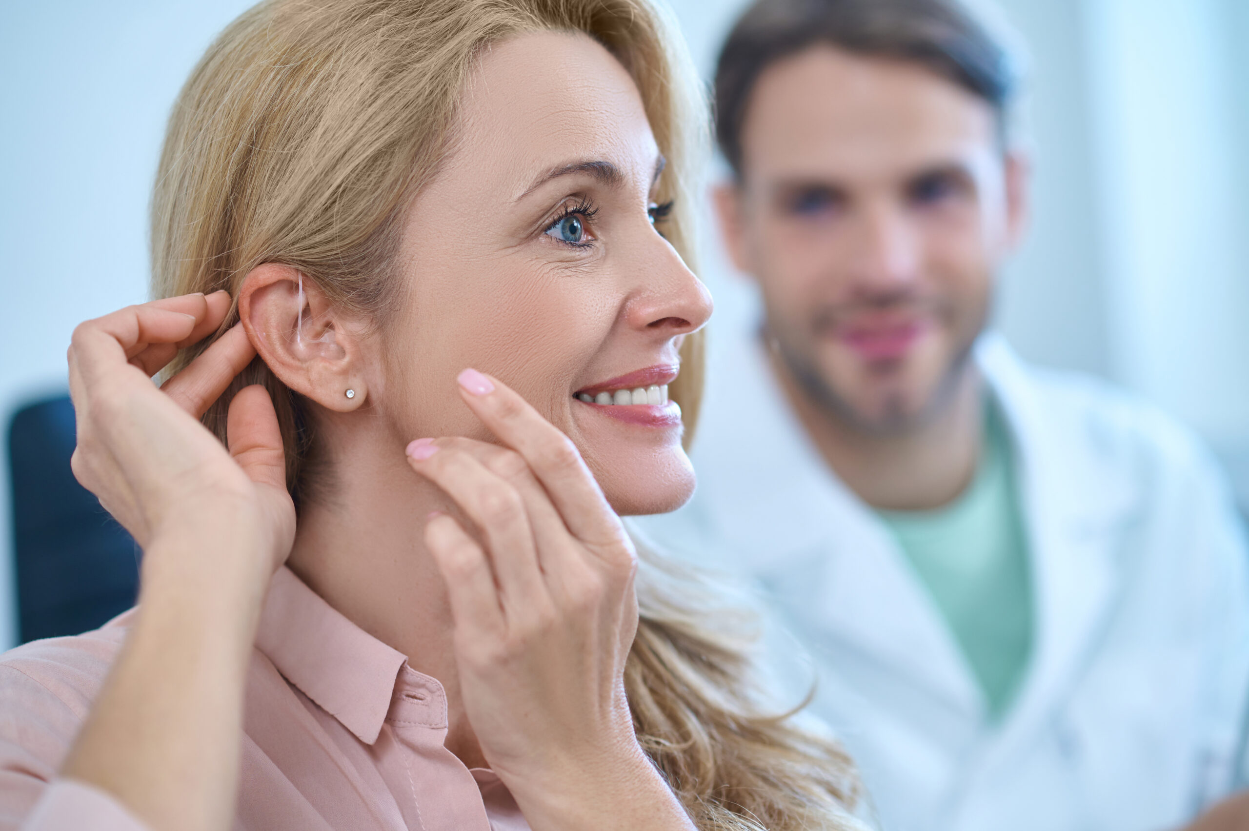 New sensation. Profile of enthusiastic pretty woman with long blond hair touching her hands to ear and man in white coat watching behind in medical office