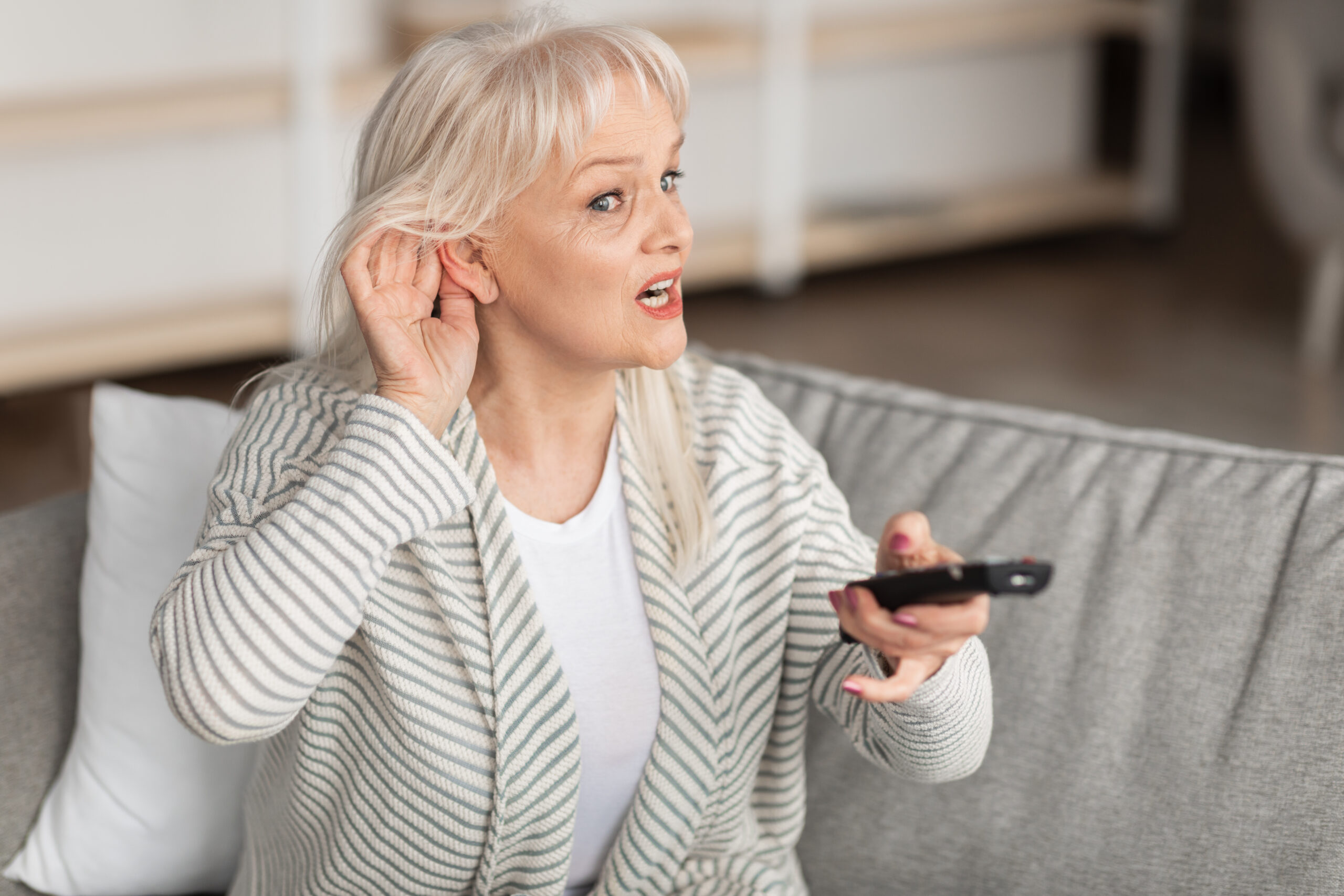 Portrait of confused senior woman with impaired hearing watching TV, trying to listen holding her hand near ear to hear better, sitting on couch in living room. Elderly lady turning volume up